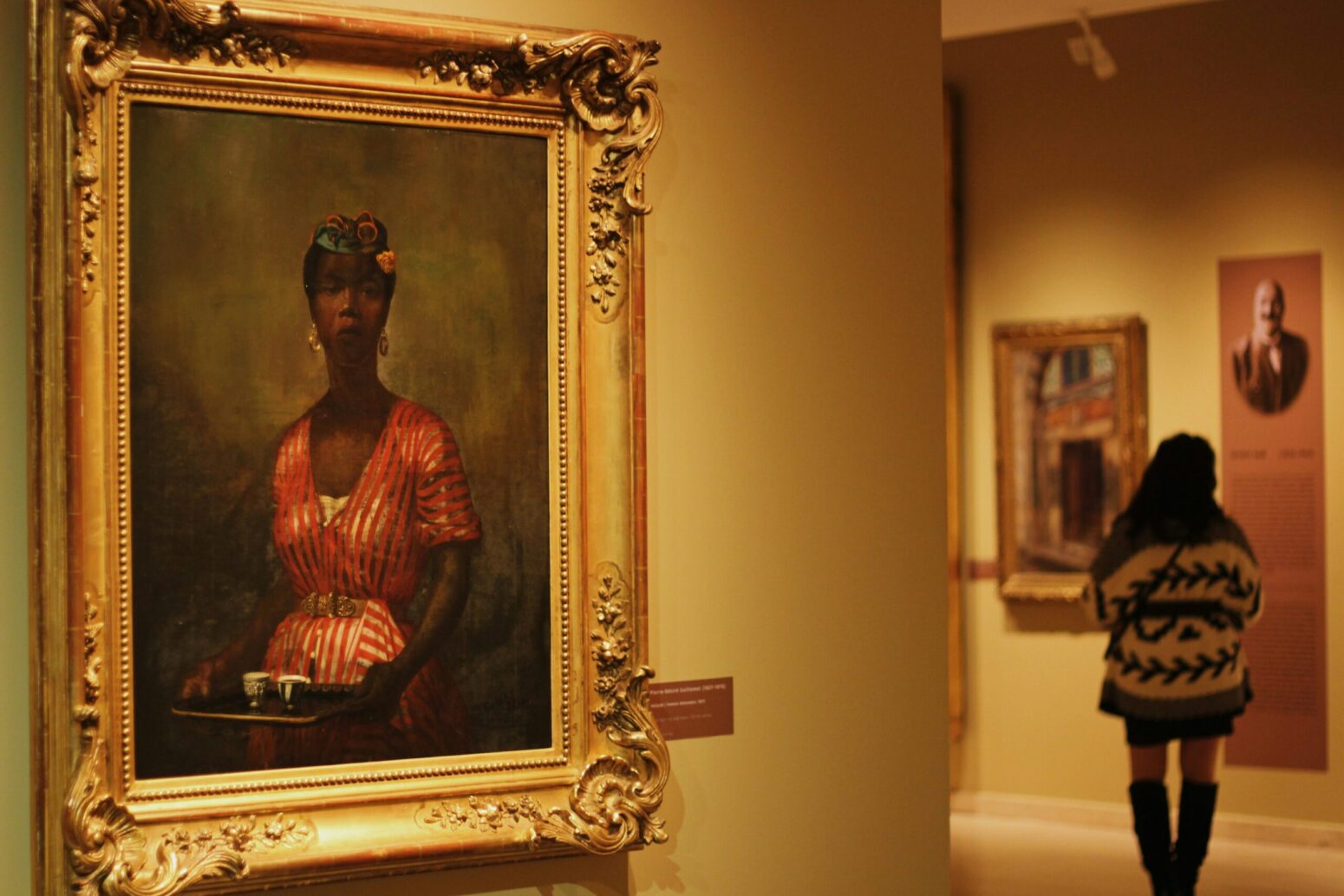 An art gallery, emphasizing the painting Halayik, or Black Servant, by Pierre-Désiré Guillemet. The painting is of a Black woman with a flowered hat and a pink-and-white-striped dress, holding a silver tray with two teacups on it.