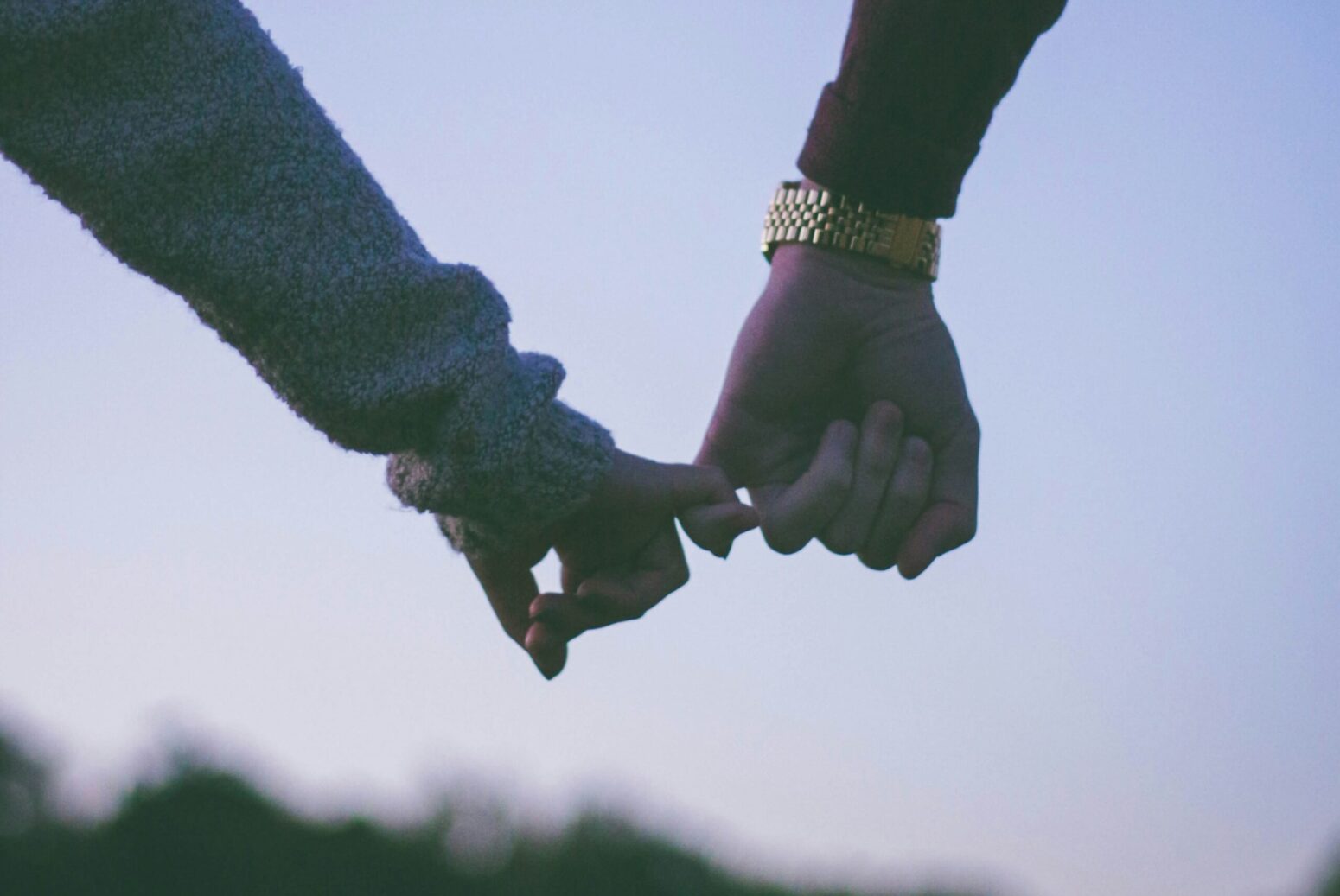 Two hands, held together by the pinky finger. One has a gold watch and a red sleeve, and the other has a blue sleeve.