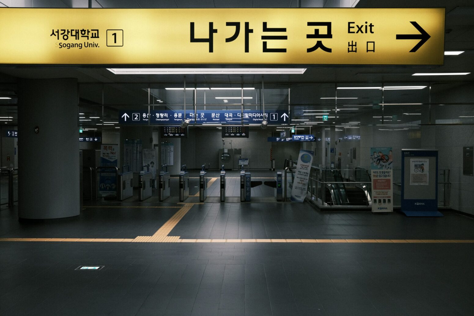 An airport terminal, with yellow signs overhead pointing to places in English, Chinese, and Korean. The airport is very dark, emphasizing the sign.