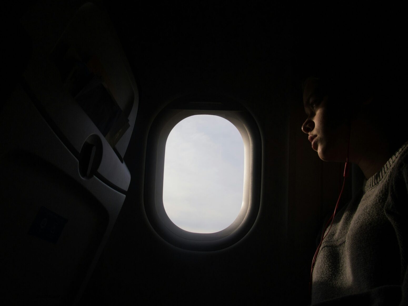 A dark passenger's seat on a plane. A window shines brightly. A woman looks out the window, seeming sad.