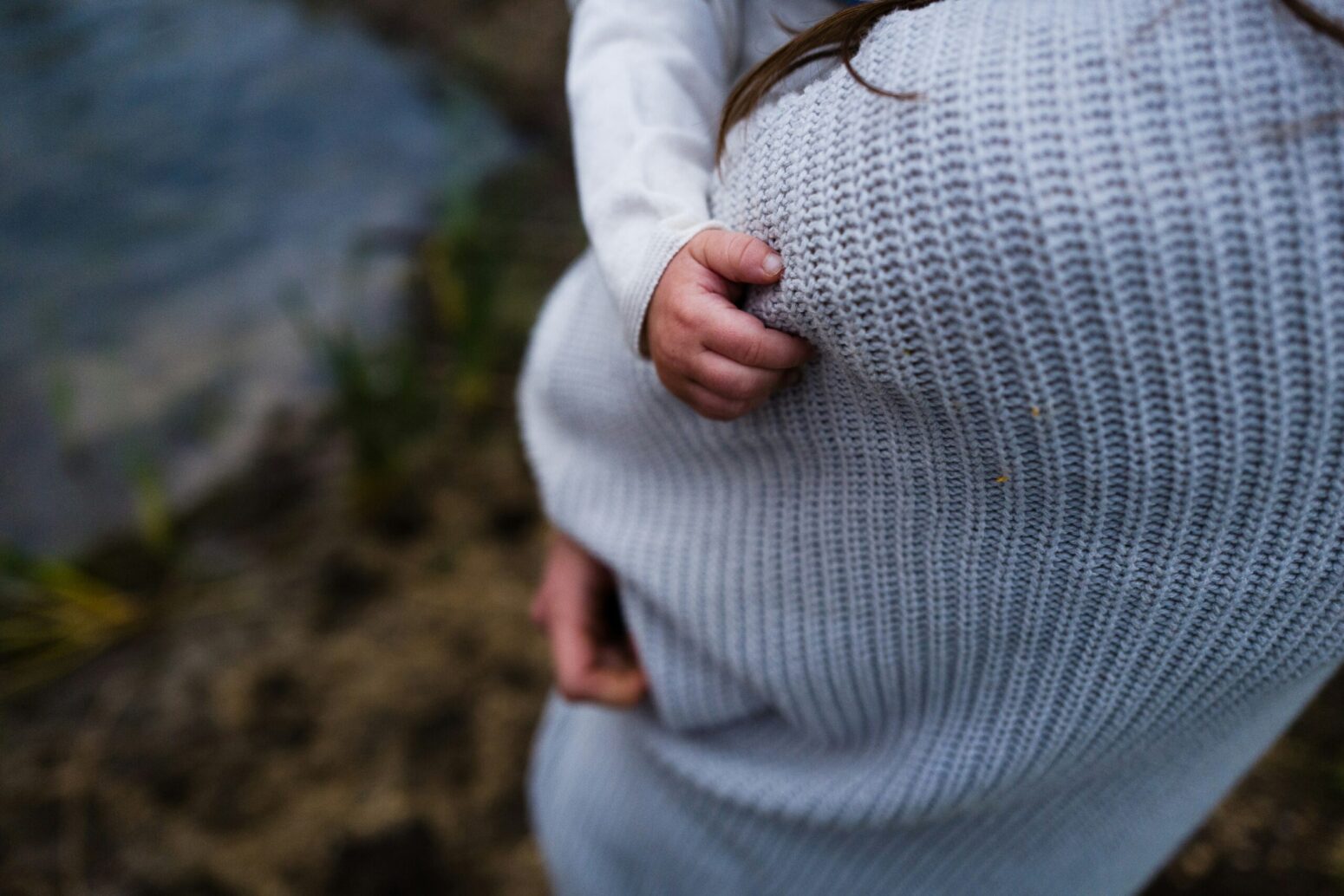 A woman in a blue sweater, overlooking the grassy shore of a lake while holding a baby.