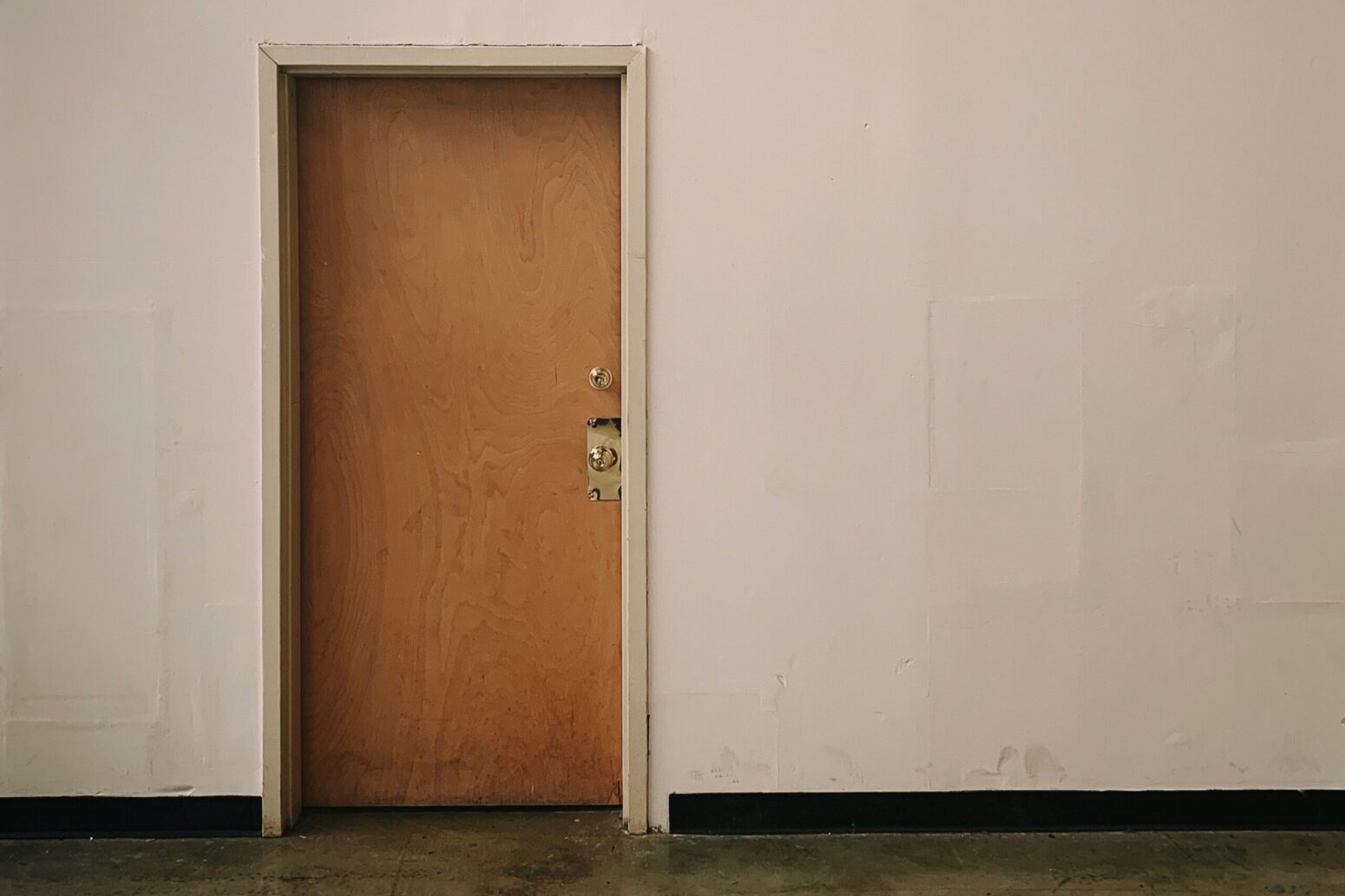 A oak door, slightly on the left of a white wall. The door has a golden handle. The floor is some sort of dark wood.
