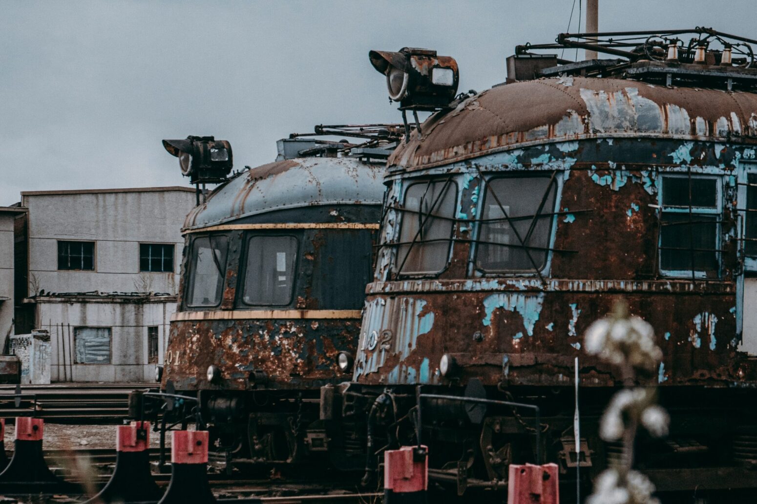 Rusted out traincars sitting in an abandoned trainyard. There are two visible, one white and one blue. A white building stands in the background.