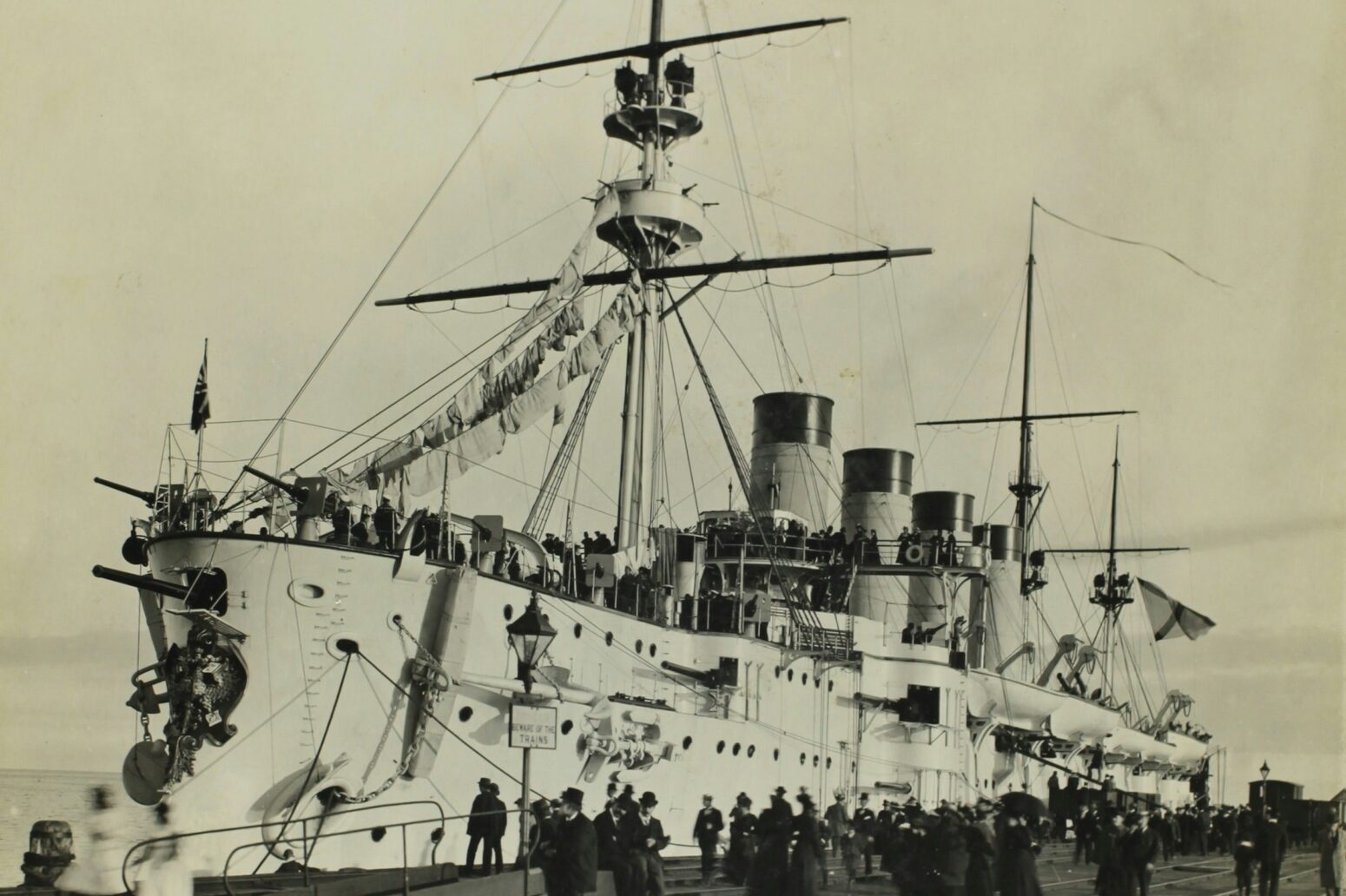A black-and-white photo of a large, old ship, docked. Many people are on the boat as well as surrounding it.