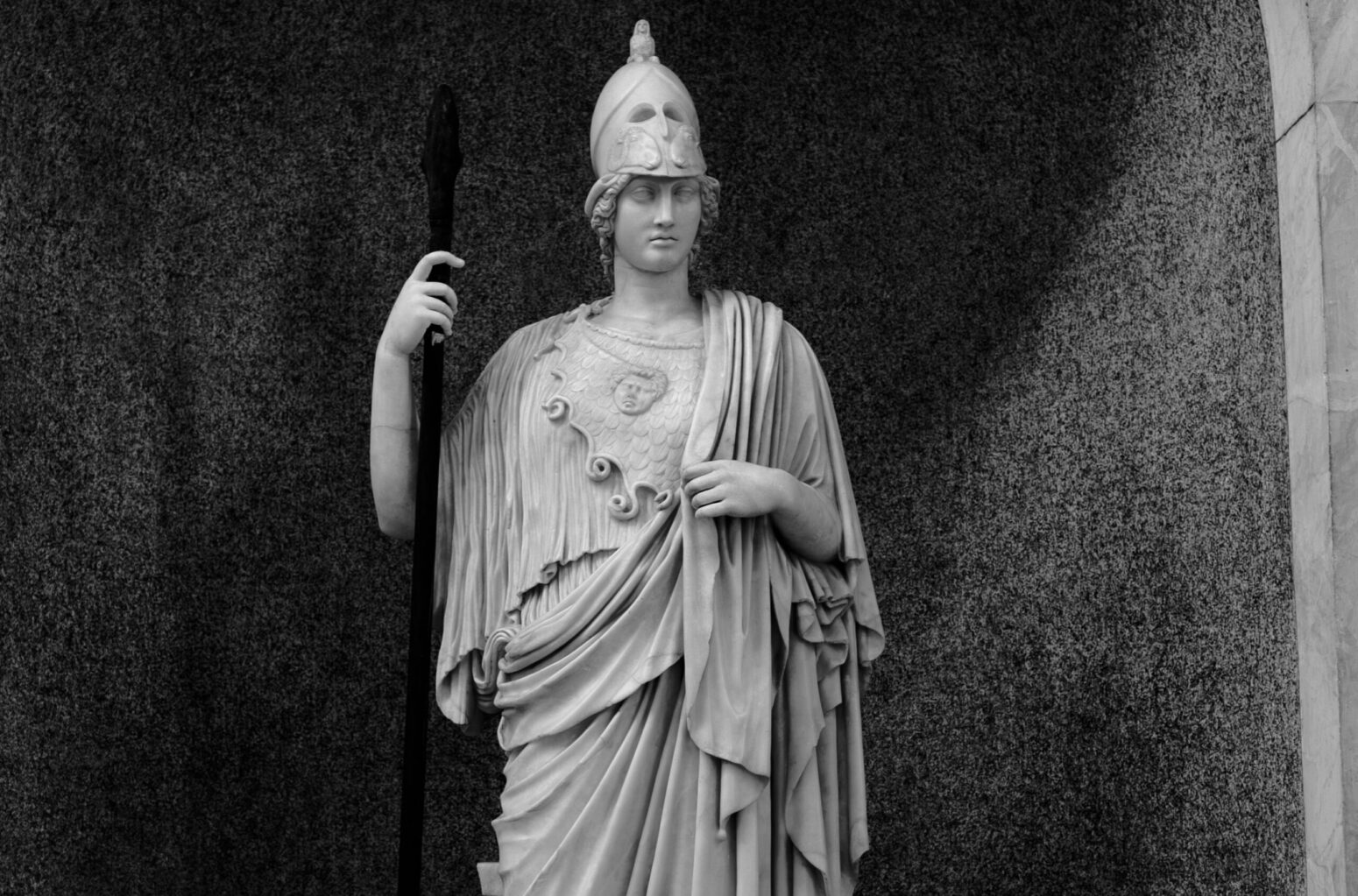A black-and-white photo of a statue of Athena in Vatican City. She wears a battle helmet on the top of her head, a draped robe, and holds a spear in her right hand.