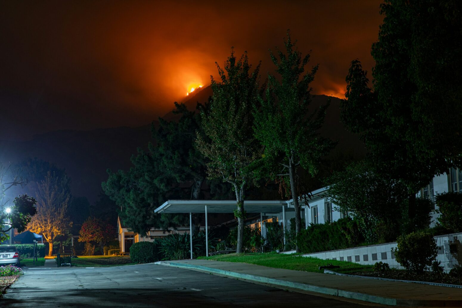 A white house on a street, surrounded by trees. A wildfire burns in the distance, behind a large mountain.