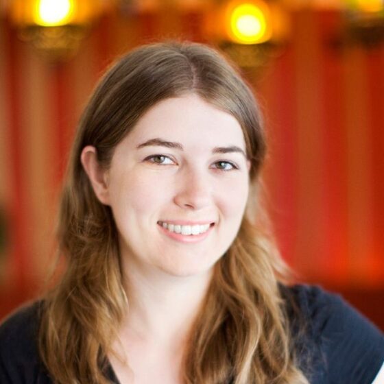 A white woman with dirty-blonde hair past her shoulders, smiling. She is standing in front of a red wall with some lights, and wearing a black, v-neck t-shirt.