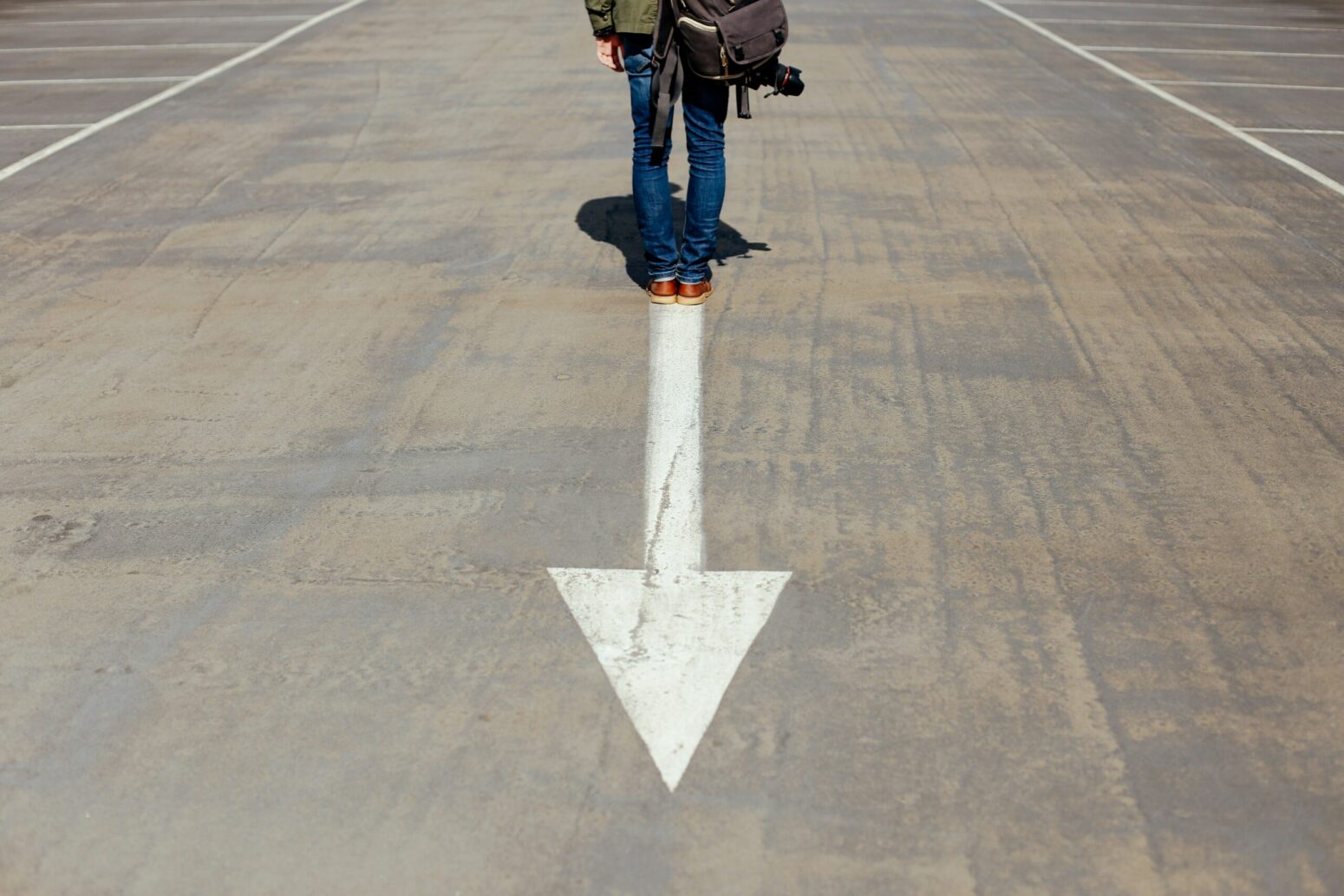 A white arrow painted on the street, pointing down. A person stands at the top of the arrow, facing the opposite direction it points. The person wears jeans, a brown jacket, and a brown-leather backpack.