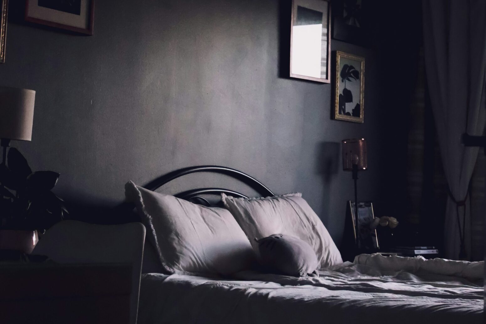 A dark bedroom with black walls. A queen-sized bed sits in the middle, with a metal bedframe and two, white pillows. Pictures hang from the wall.