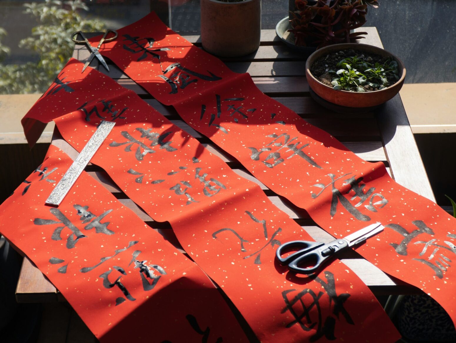 A wooden table with three red paper scrolls laid out upon it. The scrolls are adorned with calligraphy of Chinese characters. A pair of blue scissors lays on top.