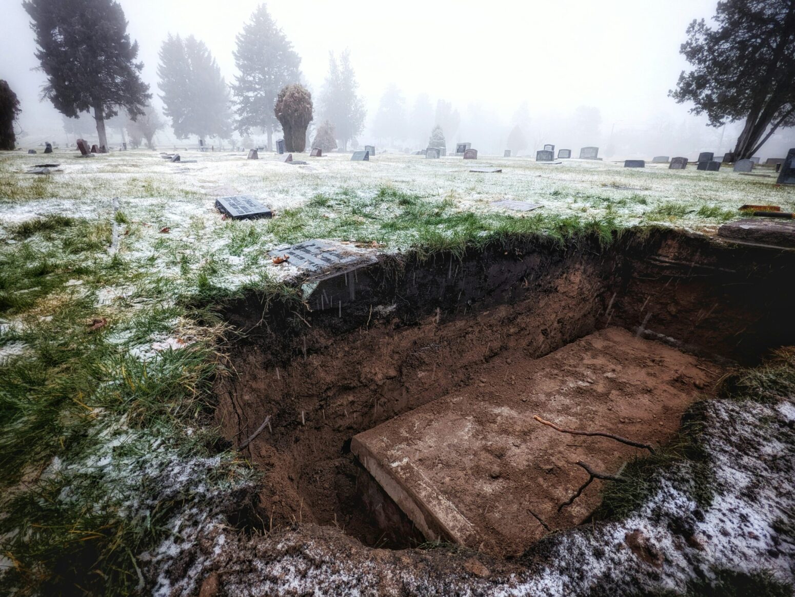 A foggy graveyard during the day. Inside a grave lies a dirty, wooden coffin.