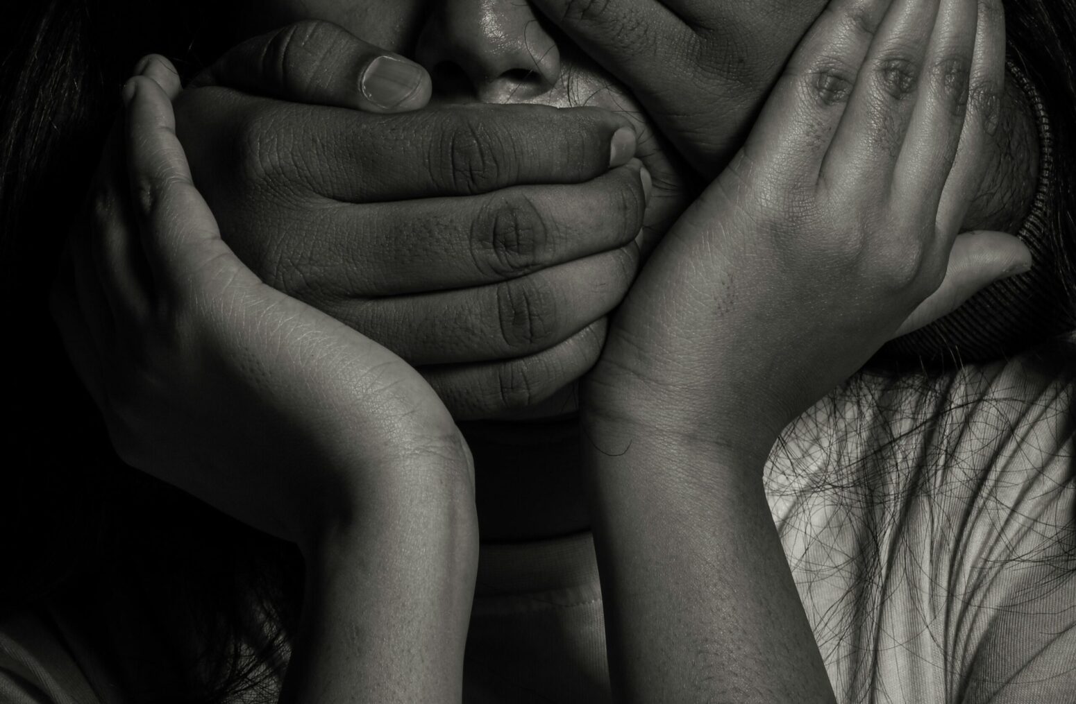 A black-and-white photo of a man's hands holding a woman by the mouth and head. The woman is crying, and holds her hands up to the man's hands.