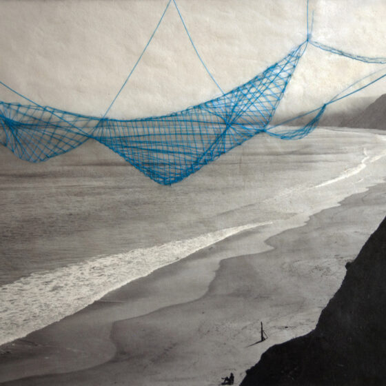 Black and white photo of a foamy wave cresting on a long, flat beach. A lone figure sits in a chair at the bottom of the image. Sewn onto the image itself is a net, made of blue thread, that appears to be suspended over the beach.