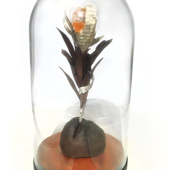 A flower is protected in a glass case shaped like a dome with a wooden bottom. The flower's petals are made of book paper, and its center is made of orange beads. The stalk and leaves of the flower are made of dried plants, and a strip of sheet music wraps around it. The flower emerges from a black piece of raw clay.