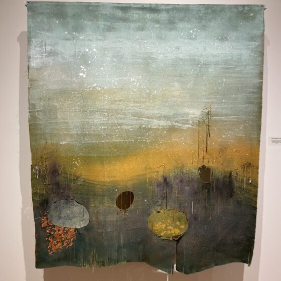 An abstract landscape on a hanging piece of canvas. The sky is blue and gray with white speckles. The horizon line fades into yellow. It looks as though there are powerlines in the distance to the right. The foreground is dark gray, with a yellow, a gray, and a black splotch at the bottom.