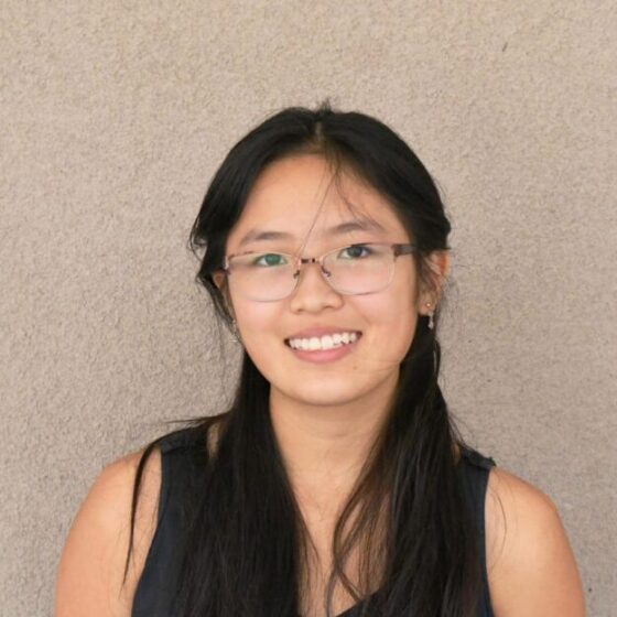 A woman with long, straight, black hair, smiling. She is standing in front of a beige wall, and wearing rectangular glasses and a black, sleeveless shirt.