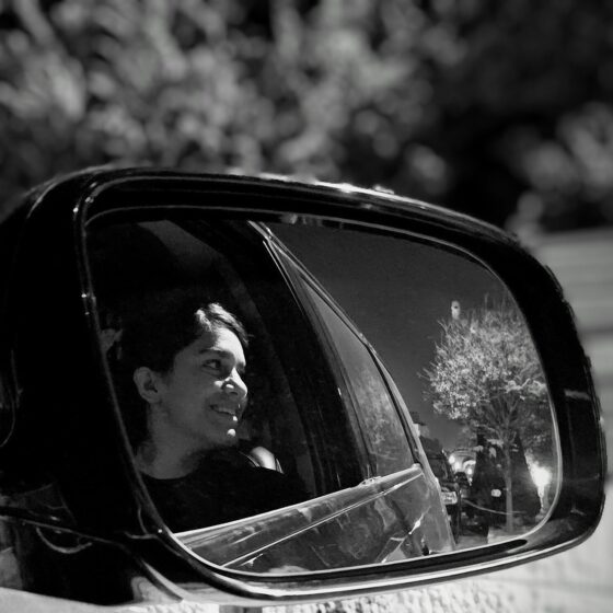 Anaya Marei smiles seated in a vehicle, her image is portrayed in the reflection of a rearview mirror. She has dark hair and has it styled as pulled back.