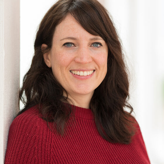 A woman with long, wavy, dark brown hair, looking forward, smiling, and leaning on a white wall to her right. She stands in front of a white background, and wears a red sweater.