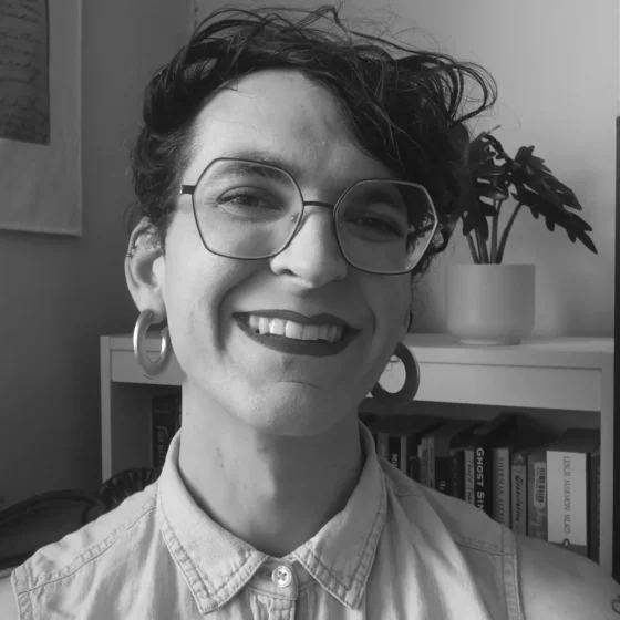 A grayscale photo of a person with short, curly hair. They are standing in front of a bookshelf with a potted plant on the top, and smiling. They are wearing a button up, hexagonal glasses, and hoop earrings.