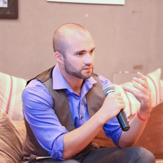 A bald man with a five o' clock shadow. He sits on a couch in a room with beige walls, holding a microphone with his right hand, and looking to the right. He wears a purple button-down with a gray vest and blue jeans.