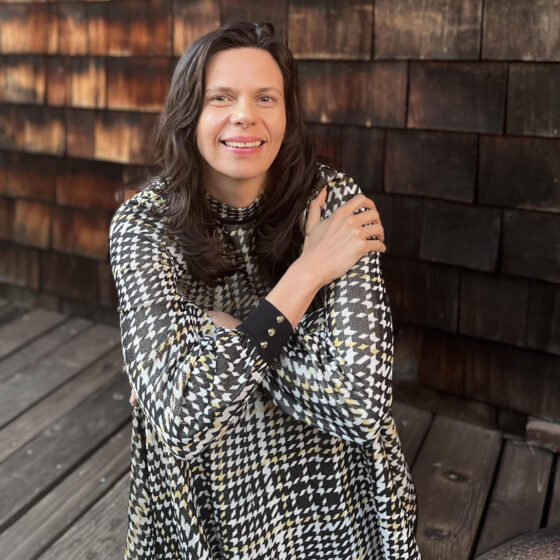 A woman with long, curly, dark hair sitting on the ground, smiling. She had her knees pulled up to her chest, and she crosses her arms over top them. She sits on a wooden floor in front of a wooden-slat wall, and wears a black-and-white plaid dress.