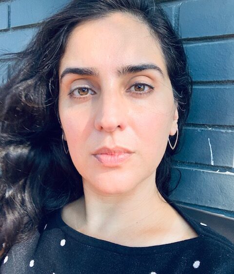 A woman with long. wavy. black hair and brown eyes. She leans up against a teal wall, and wears a black shirt with white polka dots.