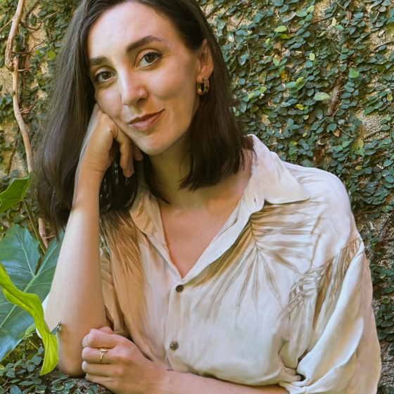 A woman with shoulder length, straight, brown hair, resting her head on her right fist. She stands in front of an ivy background, and wears a white blouse.
