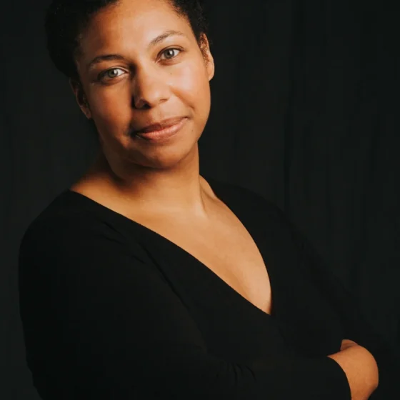 A woman with black hair back in a bun. She stands with her arms crossed, facing the right and looking at the camera. She stands in front of a black background, and wears a black, long-sleeved v-neck.
