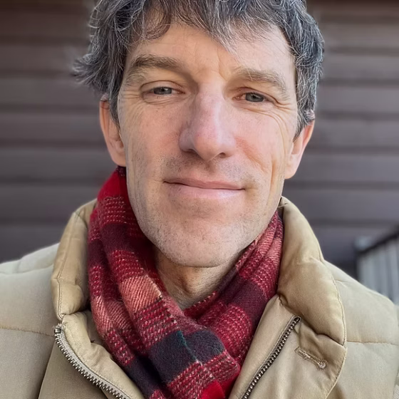 A man with short, gray hair, smiling. He stands in front of a brown house, and wears a beige coat with a red-and-black plaid scarf.