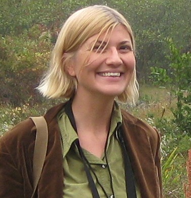 A woman with short, straight, blonde hair, smiling. She stands outside, wearing a green button-down and a brown jacket overtop.