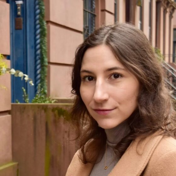 A woman with wavy, brown hair that goes just past her shoulders, smiling. She stands in front of a terracotta-colored building with blue doors, and wears a gray turtleneck with a tan coat.