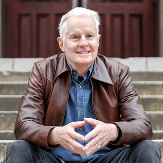 A man with white hair sitting on the stone steps leading up to a house, smiling. He has his elbows on his knees, and his fingers together. He wears dark jeans, a denim shirt, and a brown leather jacket.
