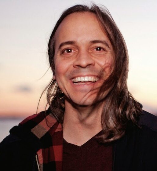 A man with straight, brown hair past his shoulders, smiling. He is standing in front of a sunset, and wearing a black shirt with a red-and-black plaid shirt overtop.