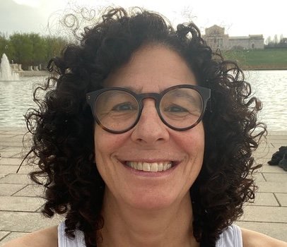A woman with black, curly hair that goes down to her shoulders, smiling. She stands outside in front of a body of water, and wears a white tank top, and black, cat-eye glasses.