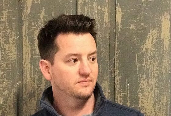 A man with short, brown hair, looking over to the right. He stands in front of a wooden background, wearing a black shirt with a collar.