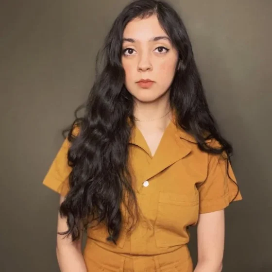 A Pakistani-Mexican woman with long, wavy, black hair. She stands in front of a gray-brown background, and wears a yellow, short-sleeved jumpsuit.