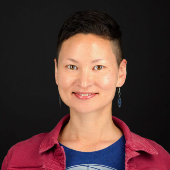 A woman with short, black hair, smiling. She stands in front of a black background, wearing a navy shirt underneath a red button-down.