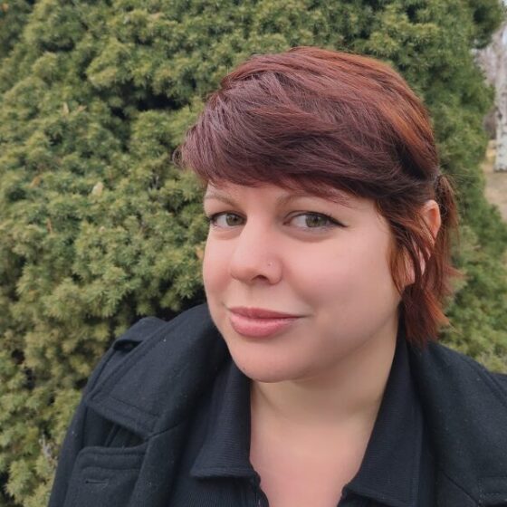 A woman with red hair, pinned back with bangs, looking at the camera and facing the left. She is standing outside in front of a tree-shrub, and wearing a black overcoat.