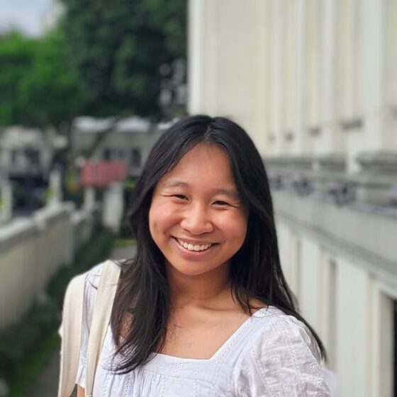 A woman with long, straight, black hair, smiling. She stands outside a building with white columns, and wears a white dress with puffy sleeves.