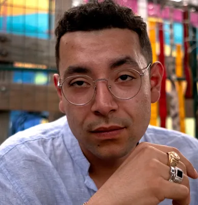 A man with short, black hair, smiling and holding his hand in near his chin. He sits in front of a blurred, colorful, building background and wears a white shirt, wire glasses, and two golden rings.