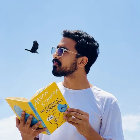 A person with black hair and a beard looking to the left. They stand in front of a sky background with a bird flying, holding a yellow book titled "The Festival of Insignificance" by Milan Kundera. They are wearing a light-blue t-shirt and square-shaped glasses.