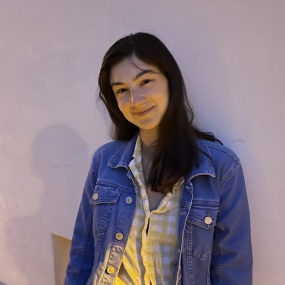 Ariel Katz smiles while leaning against a white wall, wearing a denim jacket