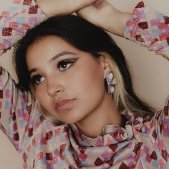 A woman with straight, black hair with blonde peekaboo-highlights. She looks over to the left, and holds her hands clasped on her head. She stands in front of a beige wall, wearing a sparkly, long-sleeved shirt with different colored squares all over it, and large, lacey earrings.