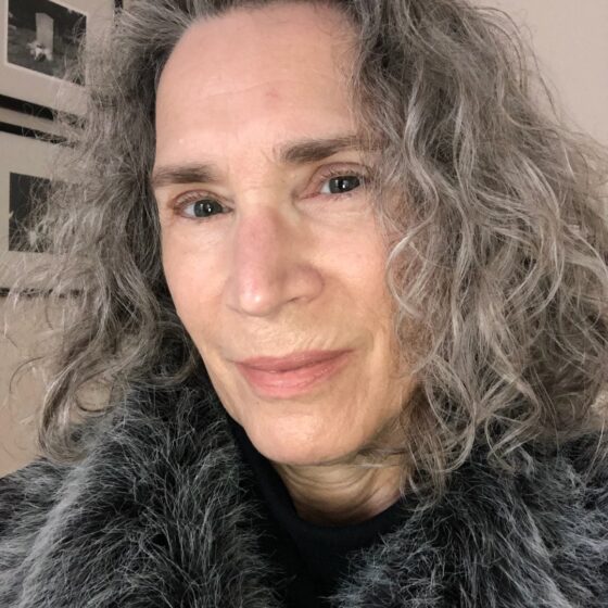 A woman with long, curly, gray hair, smiling. She is in a room with white walls and grayscale photos framed on the walls, and wears a black shirt.