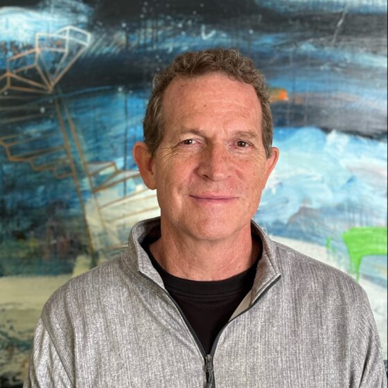 A man with short, curly, brown hair, smiling. He stands in front of an abstract art background, wearing a black shirt with a light-gray jacket over top.