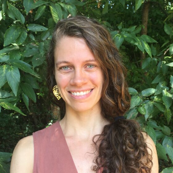 A woman with curly, brown-and-gray hair in a low, side ponytail, smiling, She stands in front of a bush, and wears a sleeveless, pink, V-neck.