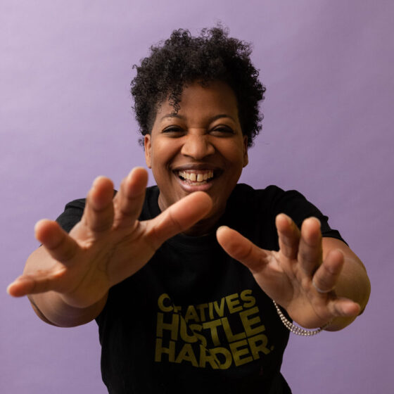 A woman with a curly, black, pixie-cut, smiling and reaching toward the camera. She stands in front of a purple background and wears a black t-shirt.