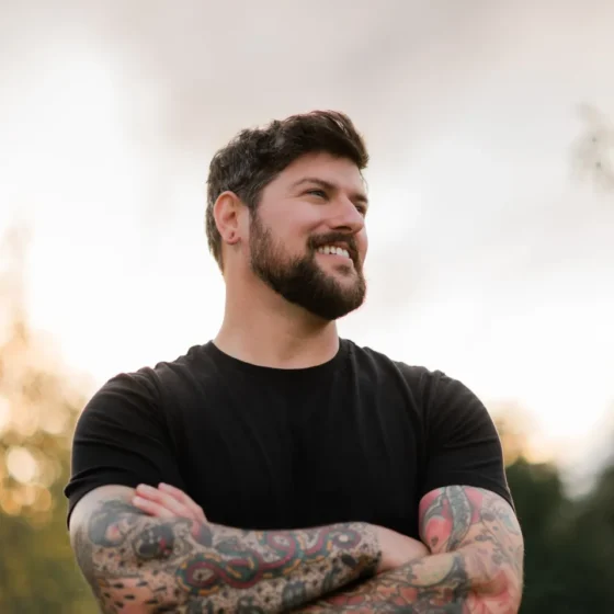 A man with short, straight, brown hair and a beard, crossing his arms, smiling, and looking to the right. He stands outside, wears a black T-shirt, and has tattoos up and down his arms.