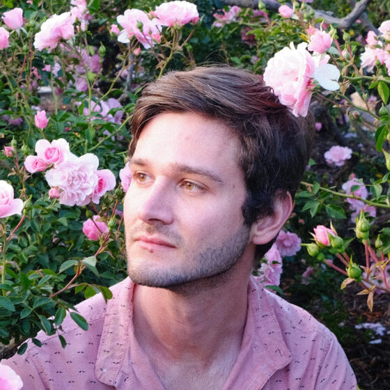 A man with short brown hair, looking to the left. He is standing amongst pink roses, and wearing a pink shirt.
