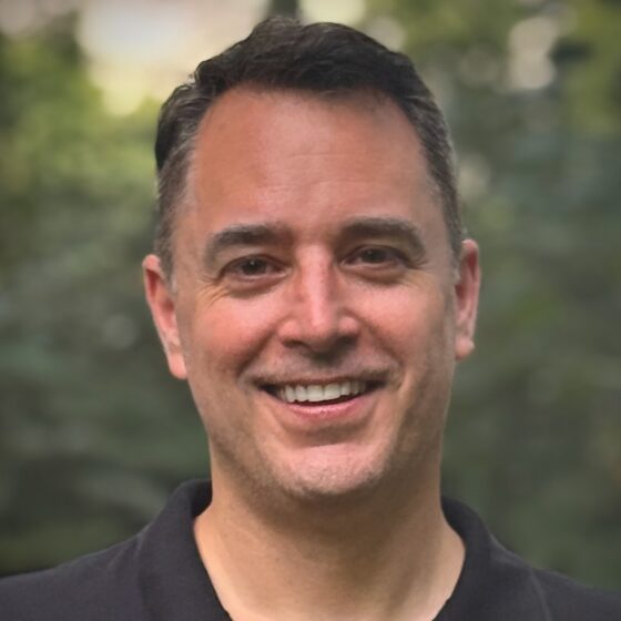Head shot of Christian Moody, a white man with dark hair that's gray at the temples. He's wearing a collared dark-gray shirt and standing outside in front of greenery